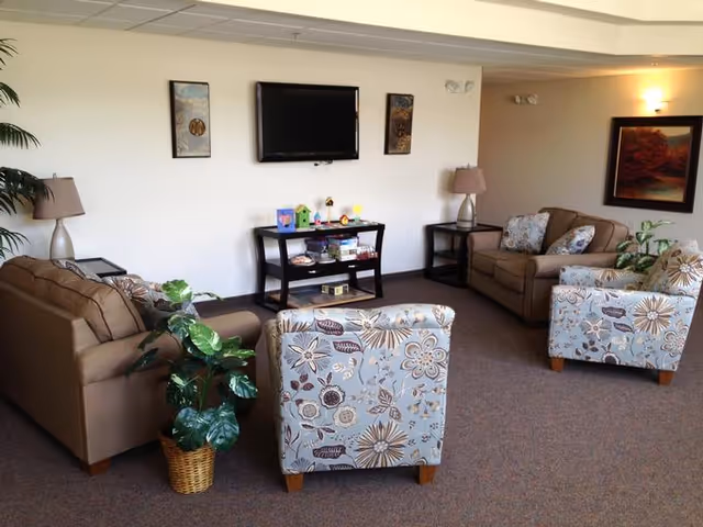 A cozy living room with two brown sofas and two floral-patterned armchairs arranged around a small black TV stand with a flat-screen TV mounted on the wall above it. The room features two table lamps on side tables, framed artwork on the walls, and a potted plant in a wicker basket on the carpeted floor.
