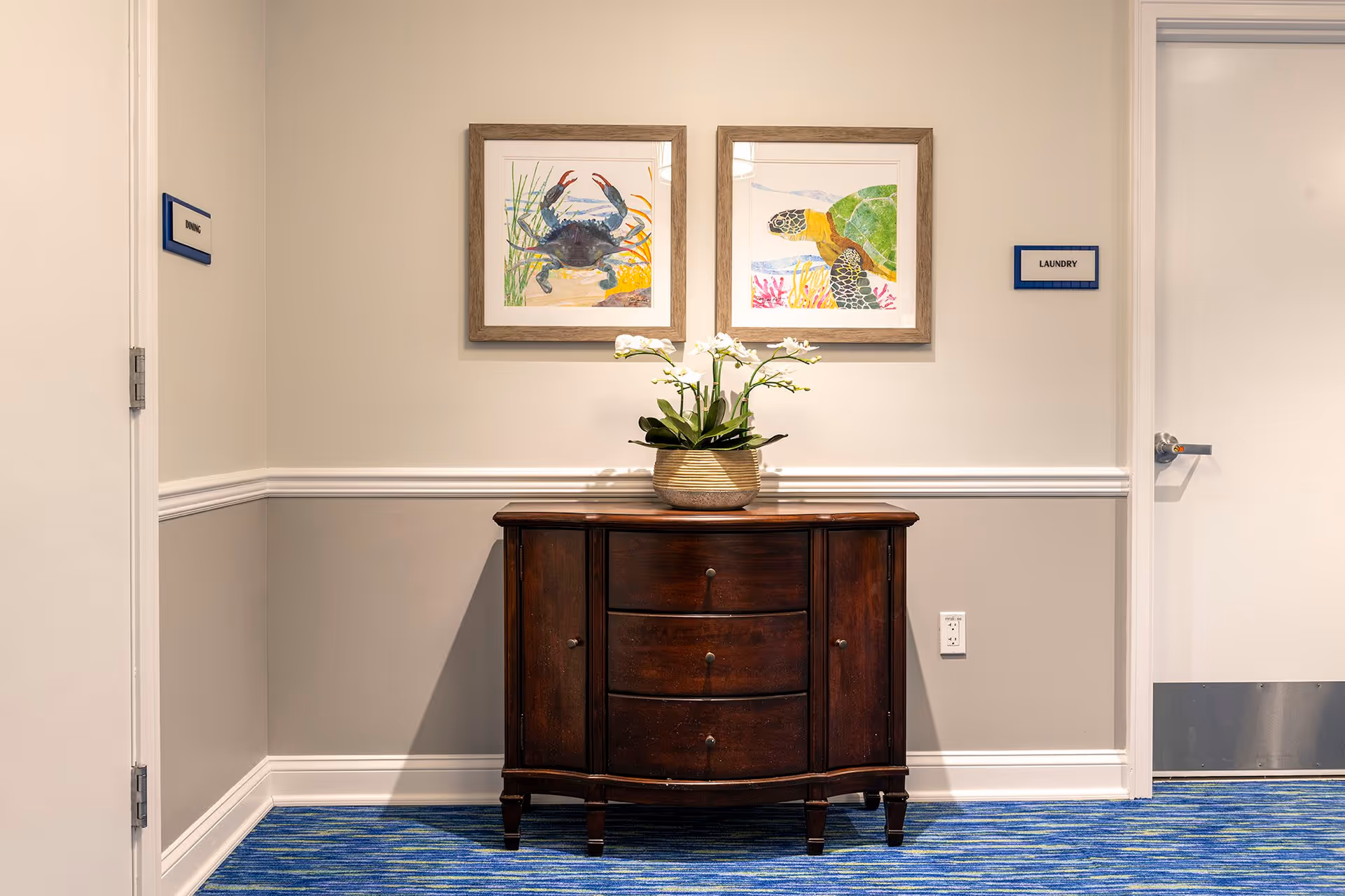 Hallway corner in a senior living facility featuring a dark wooden console table with a potted orchid beneath two framed sea-themed prints, flanked by doors labeled 'Dining' and 'Laundry'.