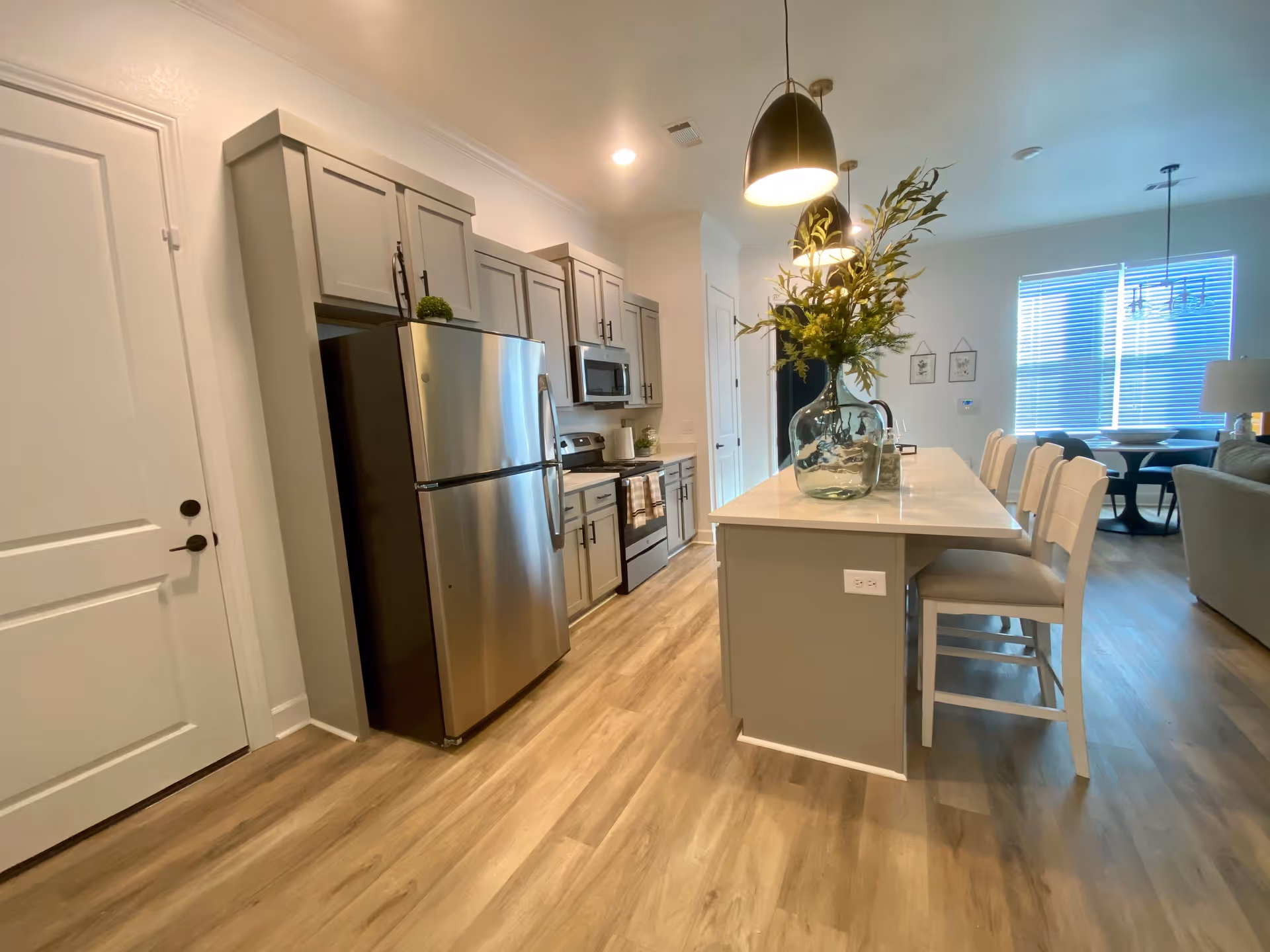 Modern kitchen area with light wood flooring, gray cabinets, stainless steel refrigerator, stove, and microwave. A large kitchen island with a white countertop has four white chairs and a glass vase with green foliage. Pendant lights hang above the island. In the background, a dining area with a round table and chairs is visible near a window with blinds.