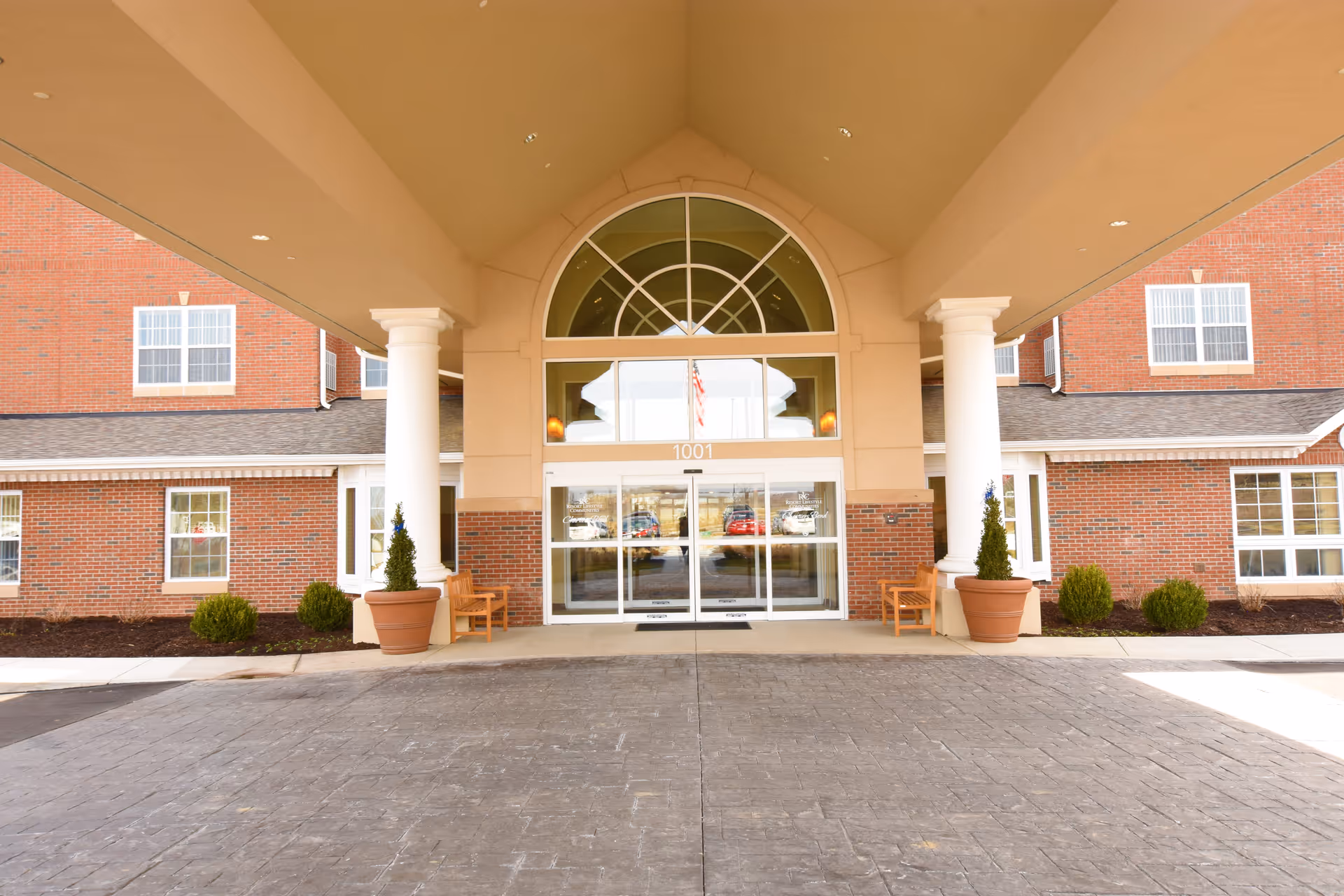 Covered main entrance of a brick retirement facility with columns, an arched window above glass doors, potted plants and benches.