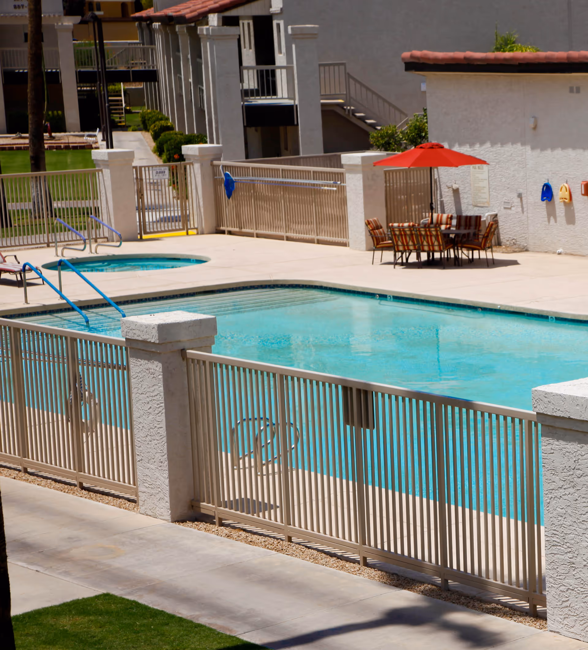 Outdoor swimming pool area at Pueblo Norte Senior Living with a fenced pool, a smaller circular hot tub, patio table with chairs under a red umbrella, and surrounding buildings.