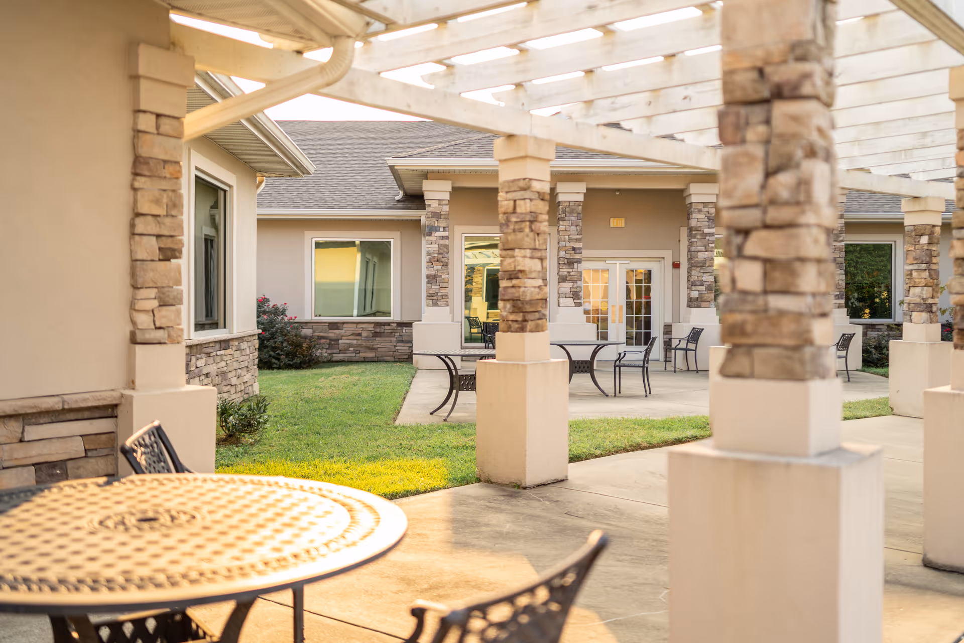 Outdoor patio area with metal tables and chairs under a pergola with stone pillars, adjacent to a building with beige walls and large windows.