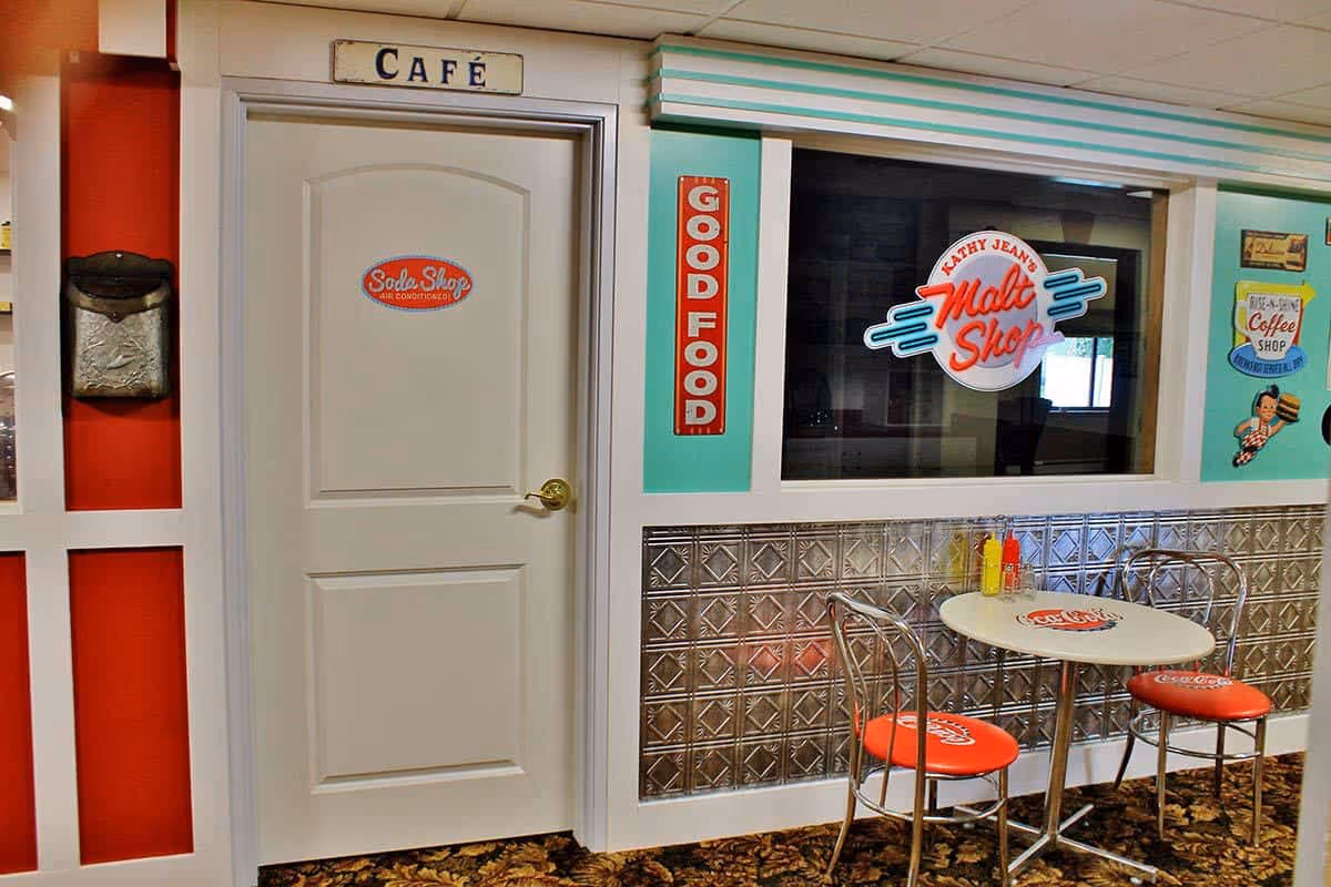 Interior view of a retro-style cafe area in a senior living facility. The scene includes a white door labeled 'Soda Shop Air Conditioned' under a sign that says 'CAFÉ'. Next to the door is a window with a sign reading 'Kathy Jean's Malt Shop' and a red vertical sign that says 'GOOD FOOD'. In front of the window is a small round table with two metal chairs that have red Coca-Cola branded seat cushions. The walls are decorated with vintage-style signs and silver paneling below the window.