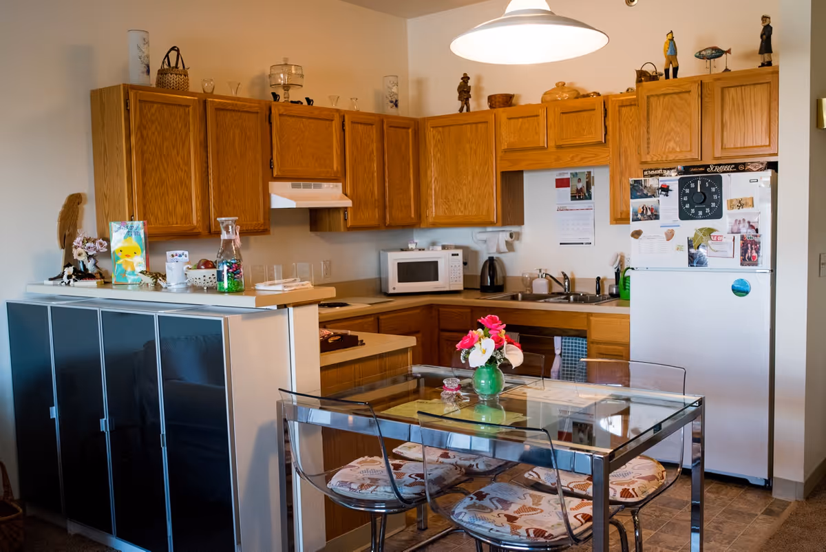 A kitchen area with wooden cabinets, a white refrigerator covered with photos and magnets, a microwave, a sink, and a glass dining table with four transparent chairs that have patterned cushions. Various decorative items and kitchen essentials are placed on the counters and cabinets.