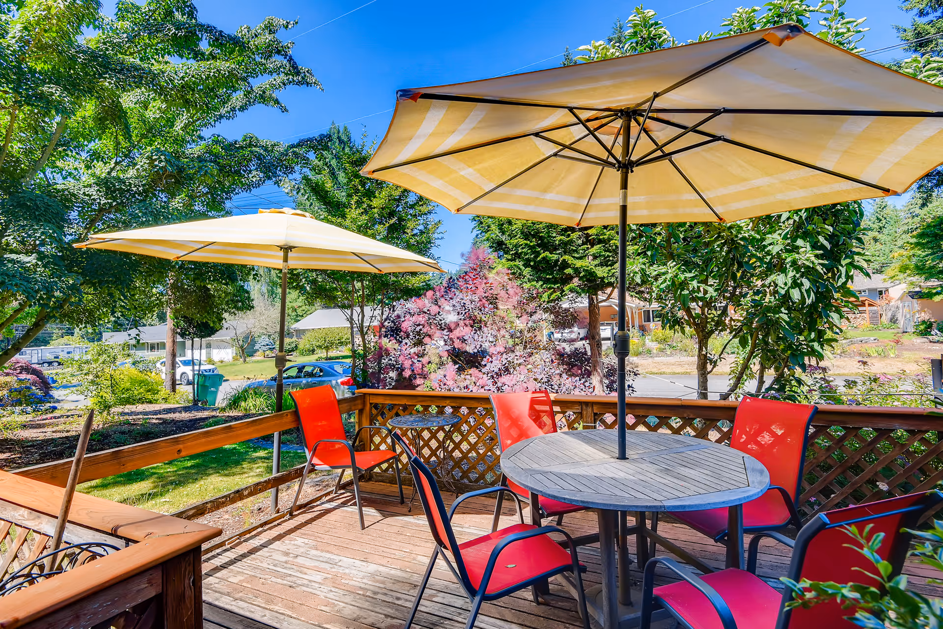 Outdoor wooden deck with a round table and six red chairs, shaded by two large yellow and white striped umbrellas. The deck is surrounded by a wooden railing with lattice panels, and there are trees, bushes, and a clear blue sky in the background.