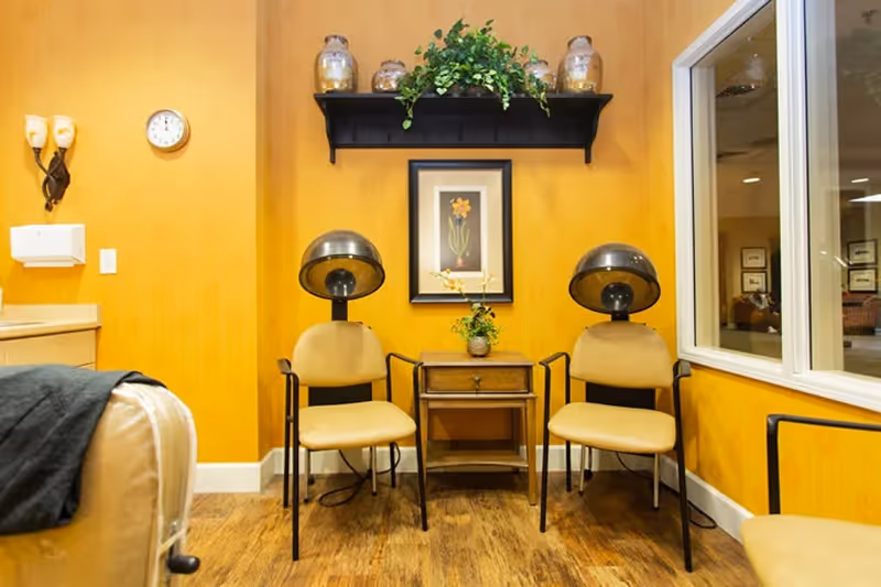Interior view of a hair salon area in a senior living facility with two vintage hair drying chairs facing forward, a small wooden table with a flower vase between them, a framed floral picture on a yellow wall, a black shelf with decorative jars and a plant above the picture, and a window showing another room.
