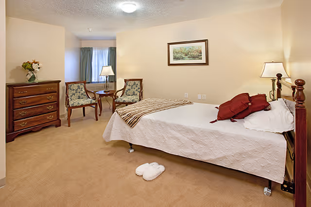 A cozy bedroom in a senior living facility featuring a single bed with white bedding and red pillows, a pair of white slippers on the carpeted floor, two floral upholstered chairs with a small round wooden table between them, a wooden dresser with a vase of flowers, a framed landscape painting on the wall, and two lamps providing warm lighting.