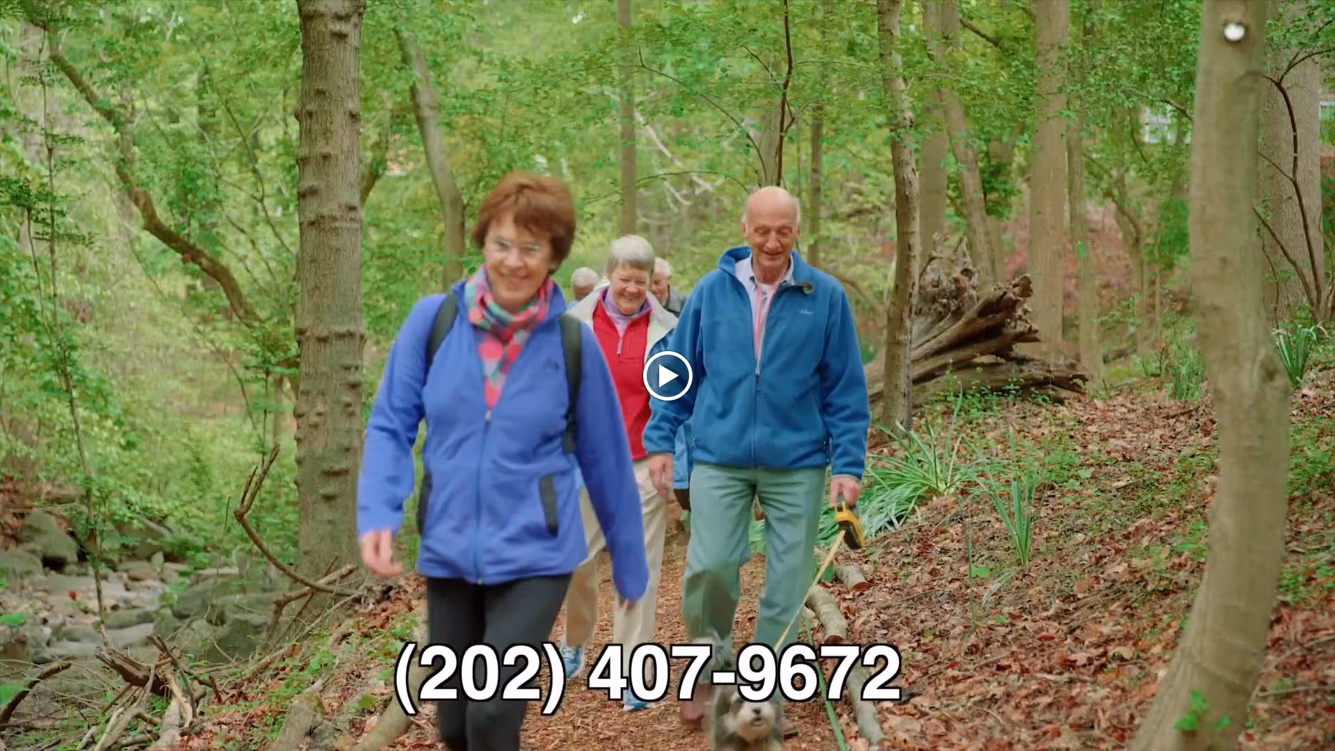 Three elderly people walking along a wooded trail with green trees and foliage around them. One woman in a blue jacket and scarf is in the foreground, followed by another woman in a red jacket and a man in a blue jacket holding a dog leash. The scene is outdoors in a natural setting.