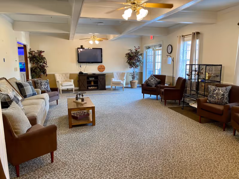 Spacious assisted living common room with sofas and armchairs arranged around a coffee table, a wall-mounted TV above a fireplace stand, and a large birdcage near the windows.