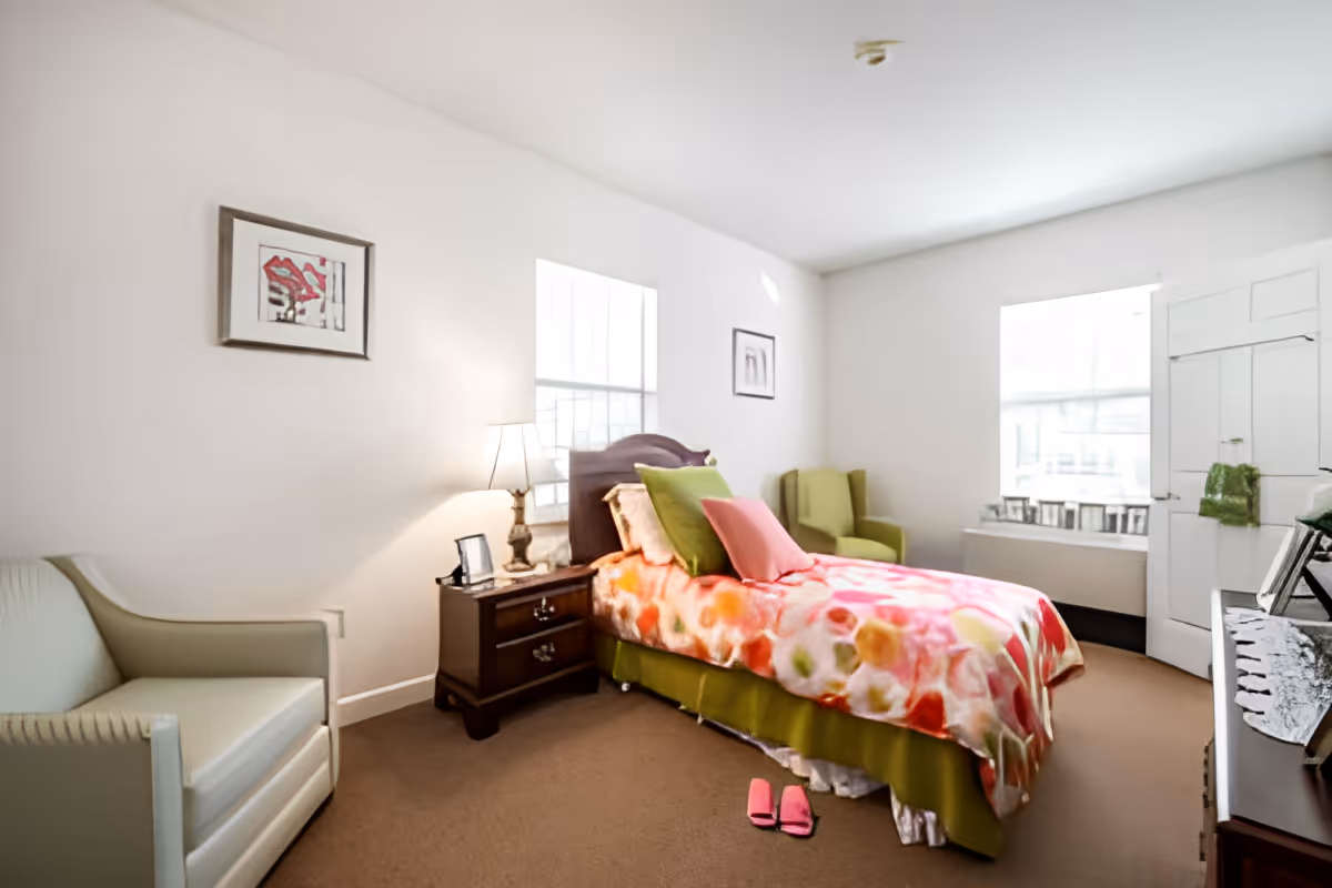 Bright furnished bedroom with a single bed covered in a colorful floral blanket, nightstand with lamp, two chairs and a pair of pink slippers on the carpet.