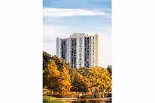 Tall light-colored apartment building rises behind autumn trees beside a reflective pond.