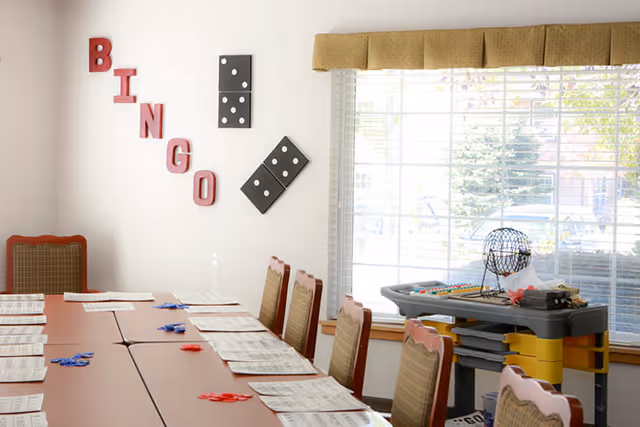 A bright activity room set up for bingo with a long table, chairs, bingo cards and markers, wall letters spelling "BINGO" and a cart with a bingo cage by the window.