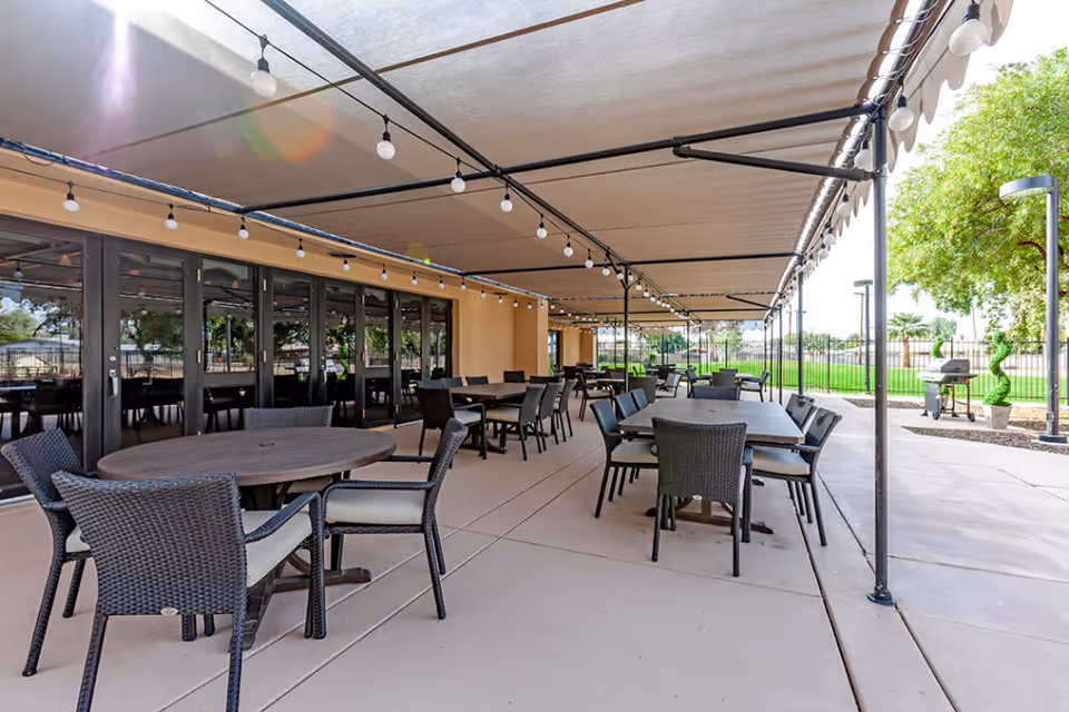 Outdoor covered patio area with multiple tables and chairs arranged for seating. The area is shaded by a large canopy with string lights hanging underneath. There is a grill and some greenery visible in the background.