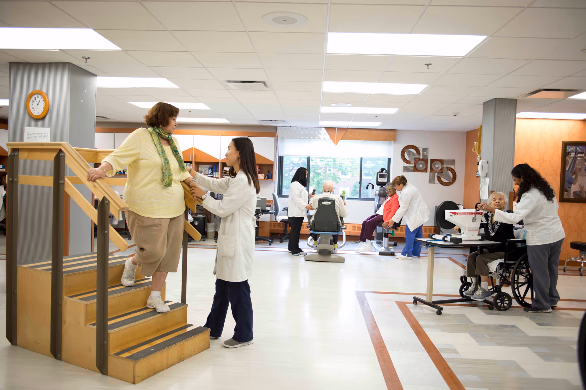 A rehabilitation center room where elderly patients are engaged in physical therapy activities. One elderly woman is walking down a small set of stairs with assistance from a healthcare worker. In the background, other elderly patients are using exercise equipment and receiving help from medical staff. The room is well-lit with large windows and has a clean, organized appearance.