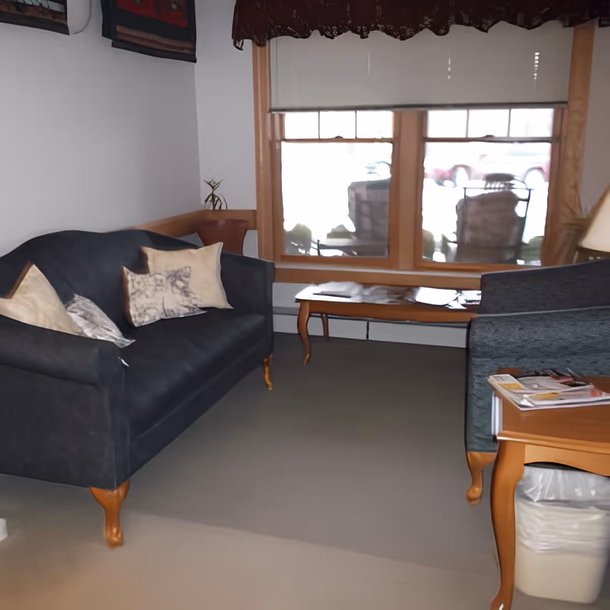 A cozy living room with two dark blue upholstered sofas adorned with decorative pillows, a wooden coffee table in front of a large window with blinds partially drawn, and a side table holding magazines and a lamp. Outside the window, patio chairs and a vehicle are visible.