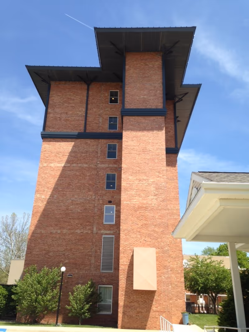 Tall multi-story red brick tower of a senior living facility with small trees and an entrance canopy under a clear blue sky.