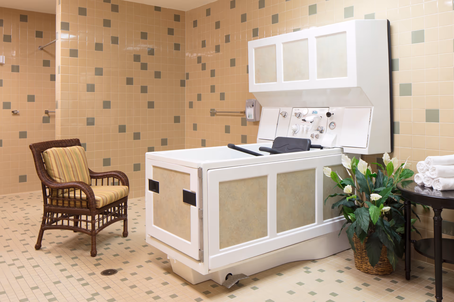 A spacious bathroom with beige and green tiled walls and floor, featuring a large white therapeutic bathtub with control knobs and a black headrest. A wicker chair with a striped cushion is placed to the left, and a small round table with rolled white towels and a potted plant with white flowers is on the right.
