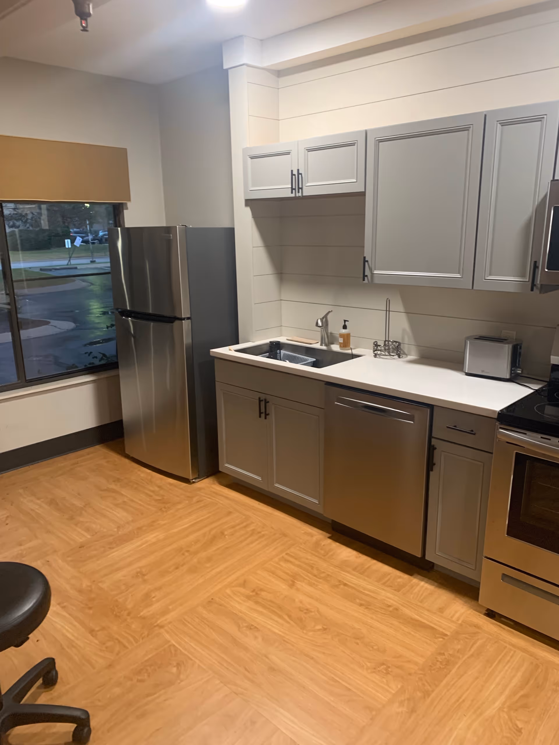 A modern kitchen area with light gray cabinets, a stainless steel refrigerator, dishwasher, and oven. There is a white countertop with a sink, a soap dispenser, and a small toaster. The floor has a wood-like finish, and there is a window with a brown shade partially covering it. A black office chair with wheels is partially visible in the lower left corner.