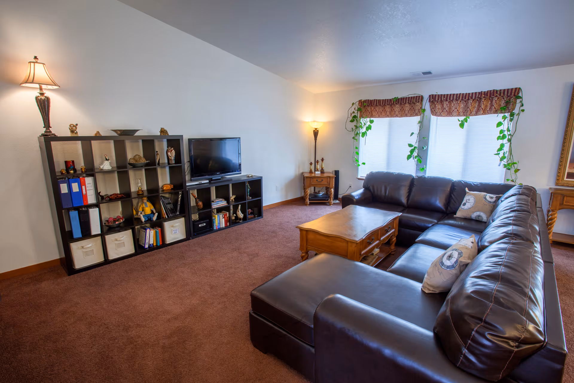A cozy living room with a large dark brown leather sectional sofa adorned with decorative pillows. There is a wooden coffee table in front of the sofa. Against the wall, there is a black shelving unit with various decorative items, books, and storage bins. A flat-screen TV is placed on the shelving unit. The room has two windows with patterned valances and trailing green plants. Two lamps provide warm lighting, one on the shelving unit and one on a small wooden side table near the windows. The floor is carpeted in a brown color.