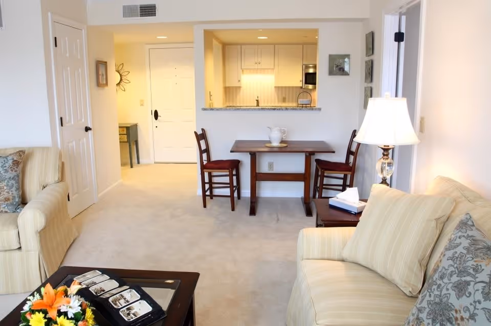 A cozy living room area with beige carpet and light-colored walls. There are two beige sofas with patterned cushions, a wooden coffee table with a flower arrangement and a tray, and a small wooden dining table with two chairs. A lamp with a white shade is on a side table next to one sofa. In the background, there is a kitchen with white cabinets and a granite countertop, and a white front door with a small table beside it.