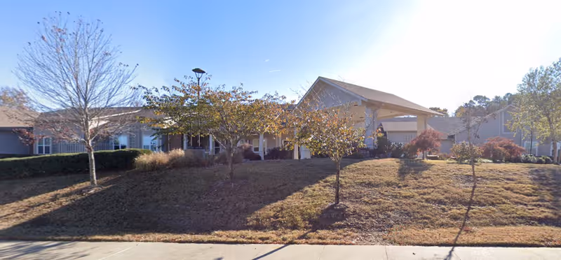 Exterior view of a single-story building with a sloped roof, surrounded by a grassy area with several small trees and shrubs. The sky is clear and sunny.
