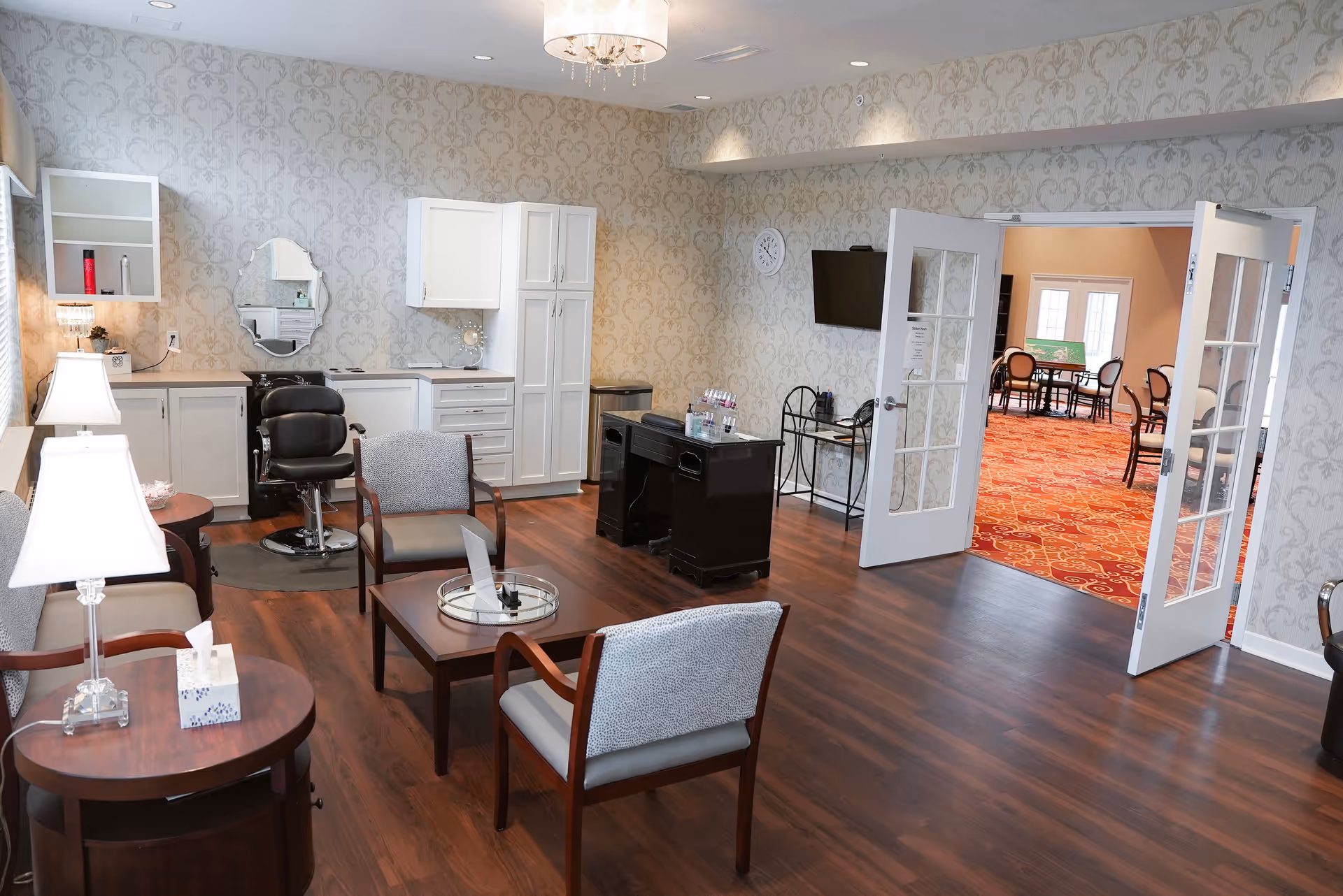 Interior room with wood flooring and patterned wallpaper, featuring a small salon area with a black salon chair, a mirror, white cabinets, and a desk with nail polish bottles. There are two upholstered chairs and a coffee table in the center, with a side table and lamp on the left. Double glass doors open to another room with red patterned carpet and several chairs around tables.