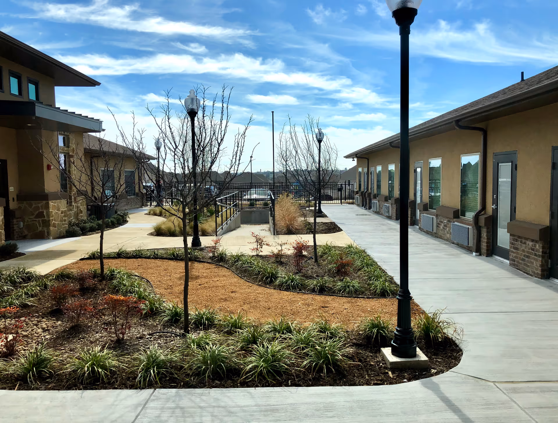 Outdoor courtyard area at Las Brisas Rehabilitation and Wellness Suites featuring a landscaped garden with small trees and plants, concrete walkways, and street lamps. The courtyard is flanked by two single-story buildings with multiple windows and doors under a partly cloudy blue sky.