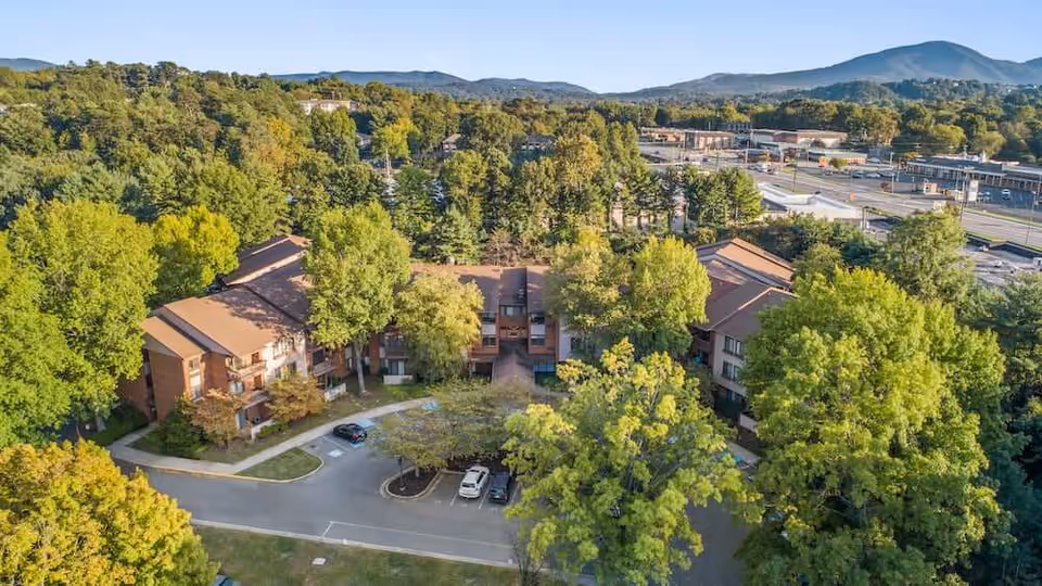 Aerial view of a multi-building senior living complex surrounded by trees, a small parking area, and distant mountains.