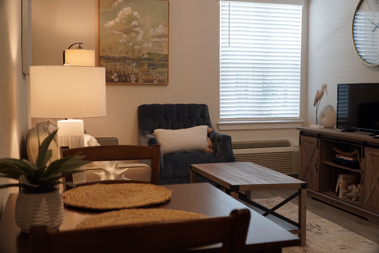 Cozy living room with a blue armchair, coffee table, TV on a wooden stand, and a dining table in the foreground.