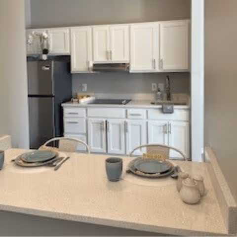 A modern kitchen area with white cabinets and drawers, a stainless steel refrigerator, and a countertop with a built-in stove. In the foreground, there is a dining counter set with two place settings including plates, bowls, cups, and utensils, along with a small teapot and creamer.