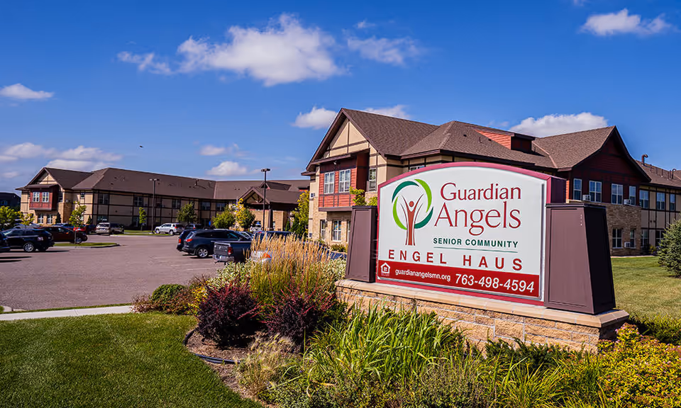 Exterior view of Guardian Angels Engel Haus Senior Living facility with a large sign in the foreground displaying the facility name, logo, website, and phone number. The building is two stories with a mix of brick and siding, surrounded by a parking lot, green grass, and landscaping under a blue sky with some clouds.