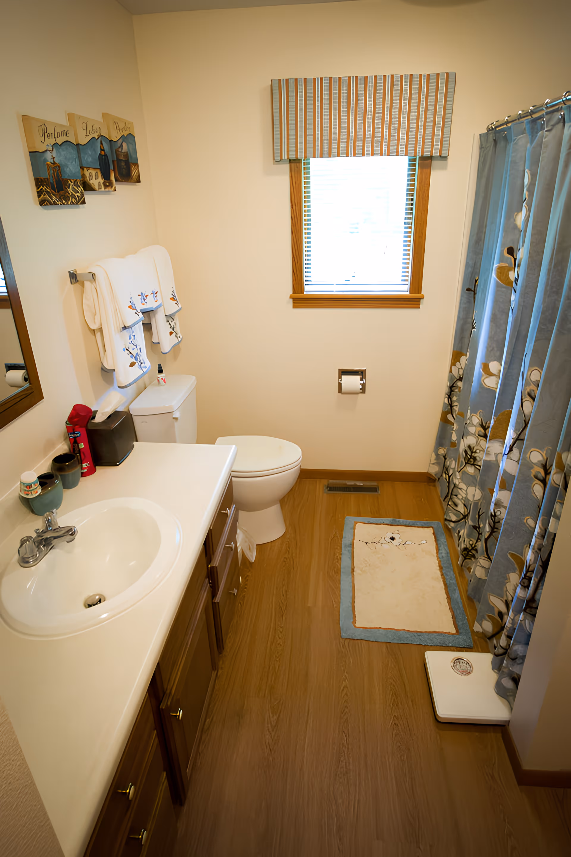 A clean bathroom with a white sink and countertop on the left, wooden cabinets below, a toilet next to the sink, and a window with blinds and a striped valance above the toilet. There are decorative towels hanging on a rack above the toilet, a floral shower curtain on the right, a small rug on the wooden floor, and a white scale near the shower.
