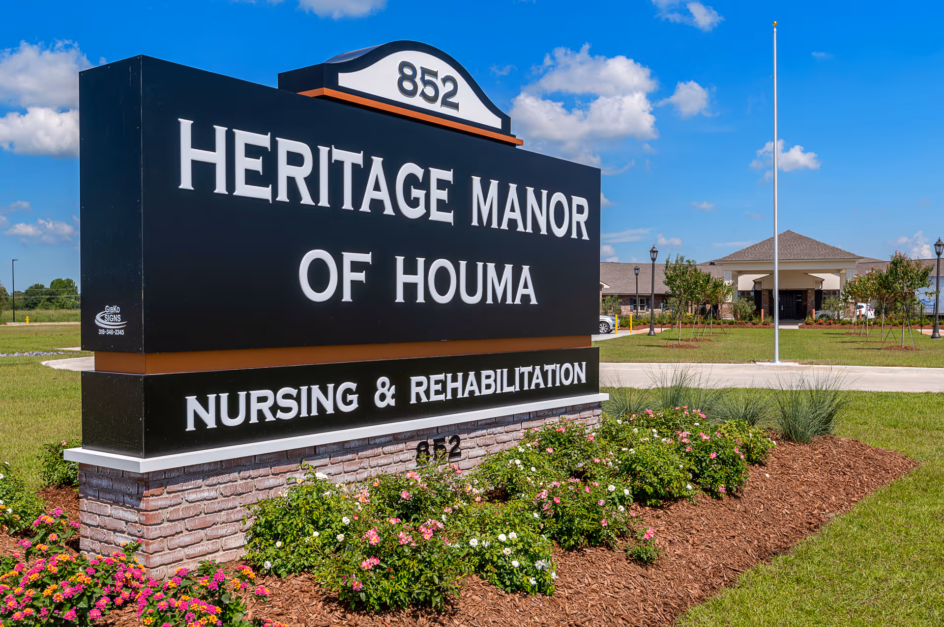 Large black sign reading "Heritage Manor of Houma Nursing & Rehabilitation" on a landscaped lawn with the facility building and blue sky in the background.