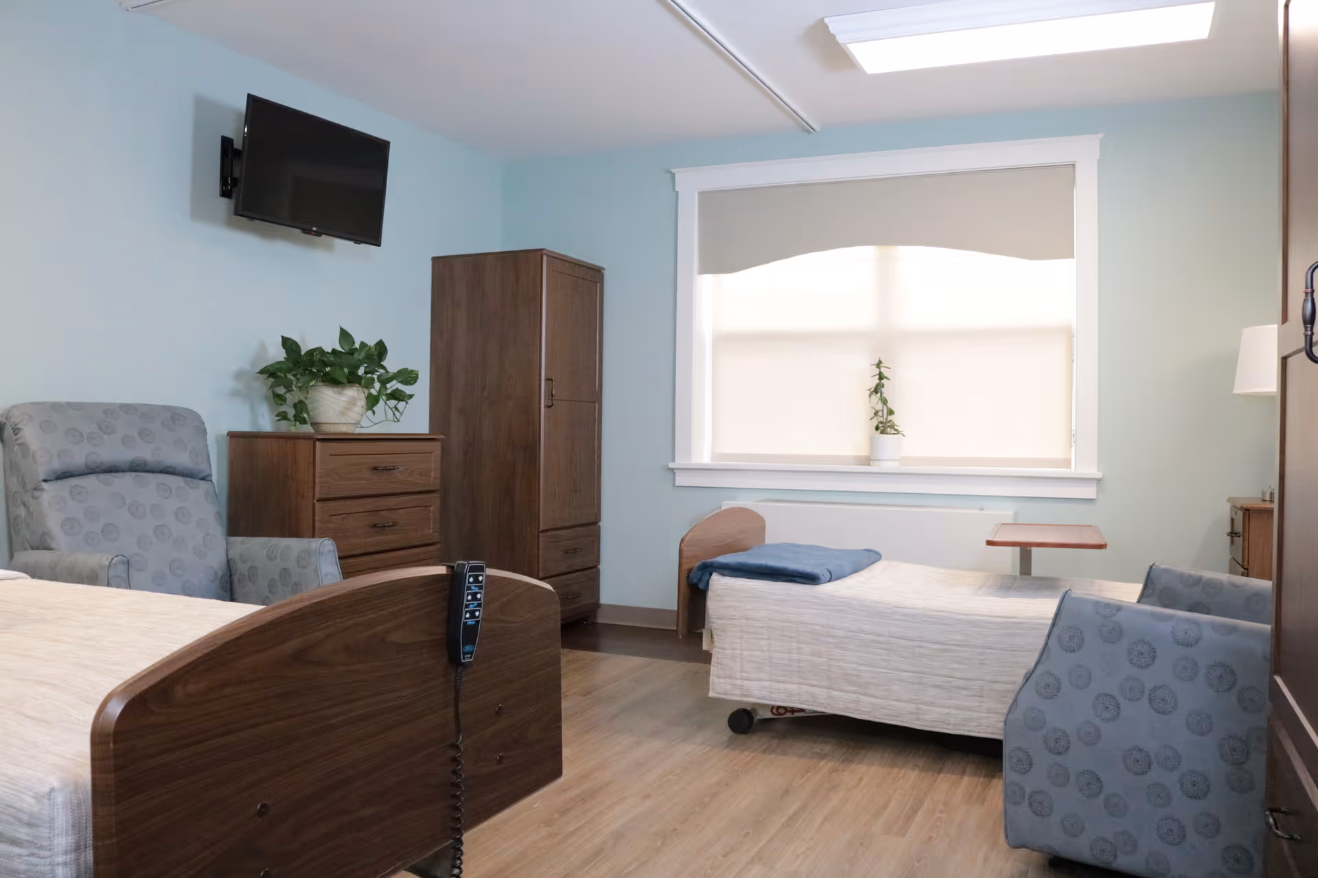 A tidy senior living bedroom with two beds, armchairs, a wall-mounted TV, and wooden furniture beneath a large window.