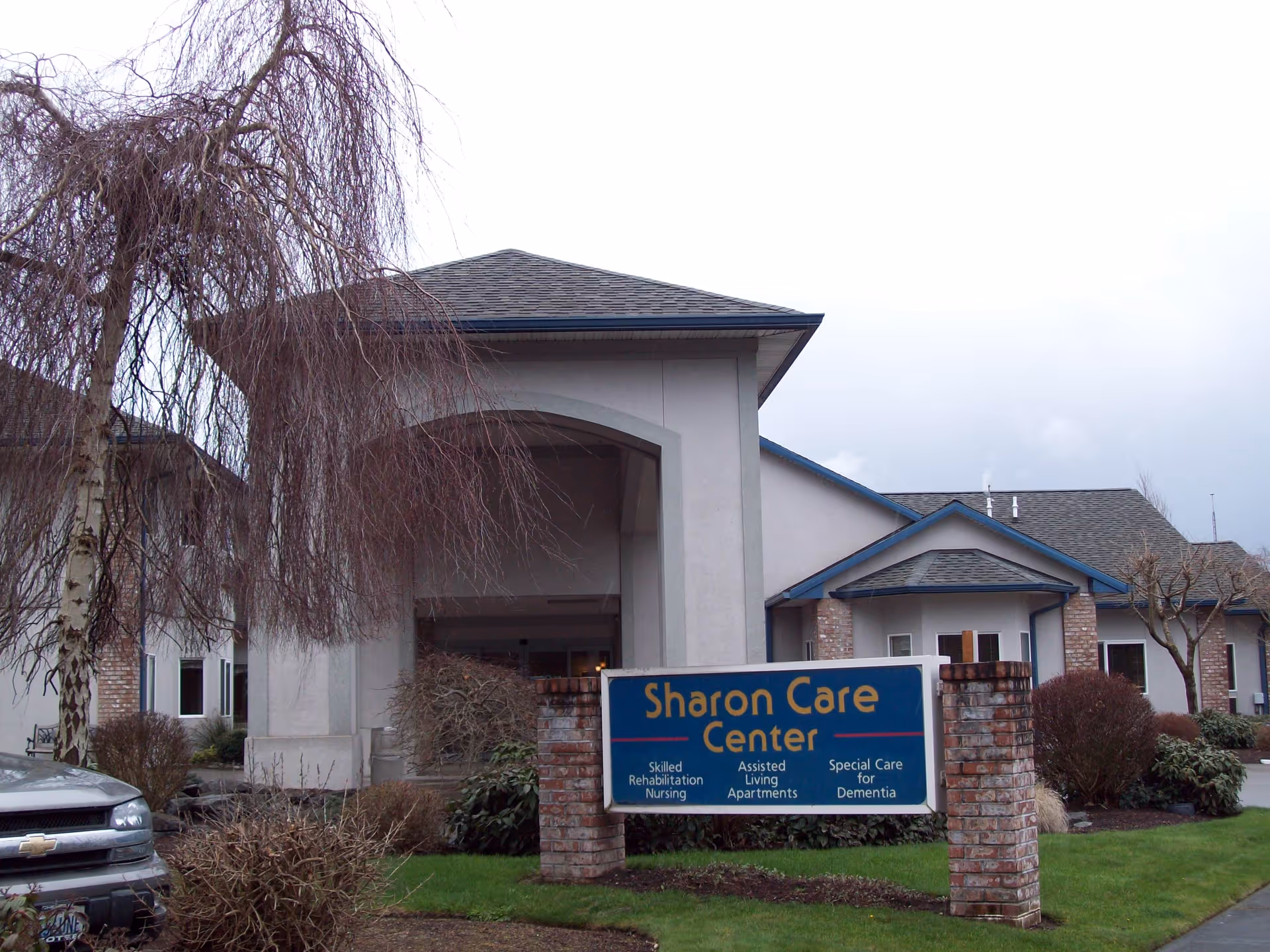 Exterior view of Sharon Care Center building with a sign in front displaying services including Skilled Rehabilitation Nursing, Assisted Living Apartments, and Special Care for Dementia. The building has a covered entrance, brick pillars, and surrounding landscaping with trees and bushes.