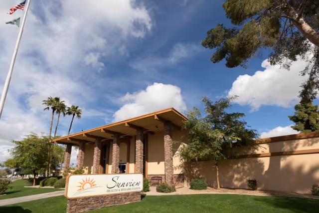 Exterior view of Sunview Health and Rehabilitation Center building with stone pillars, a sign in front, green lawn, trees, and a partly cloudy sky.