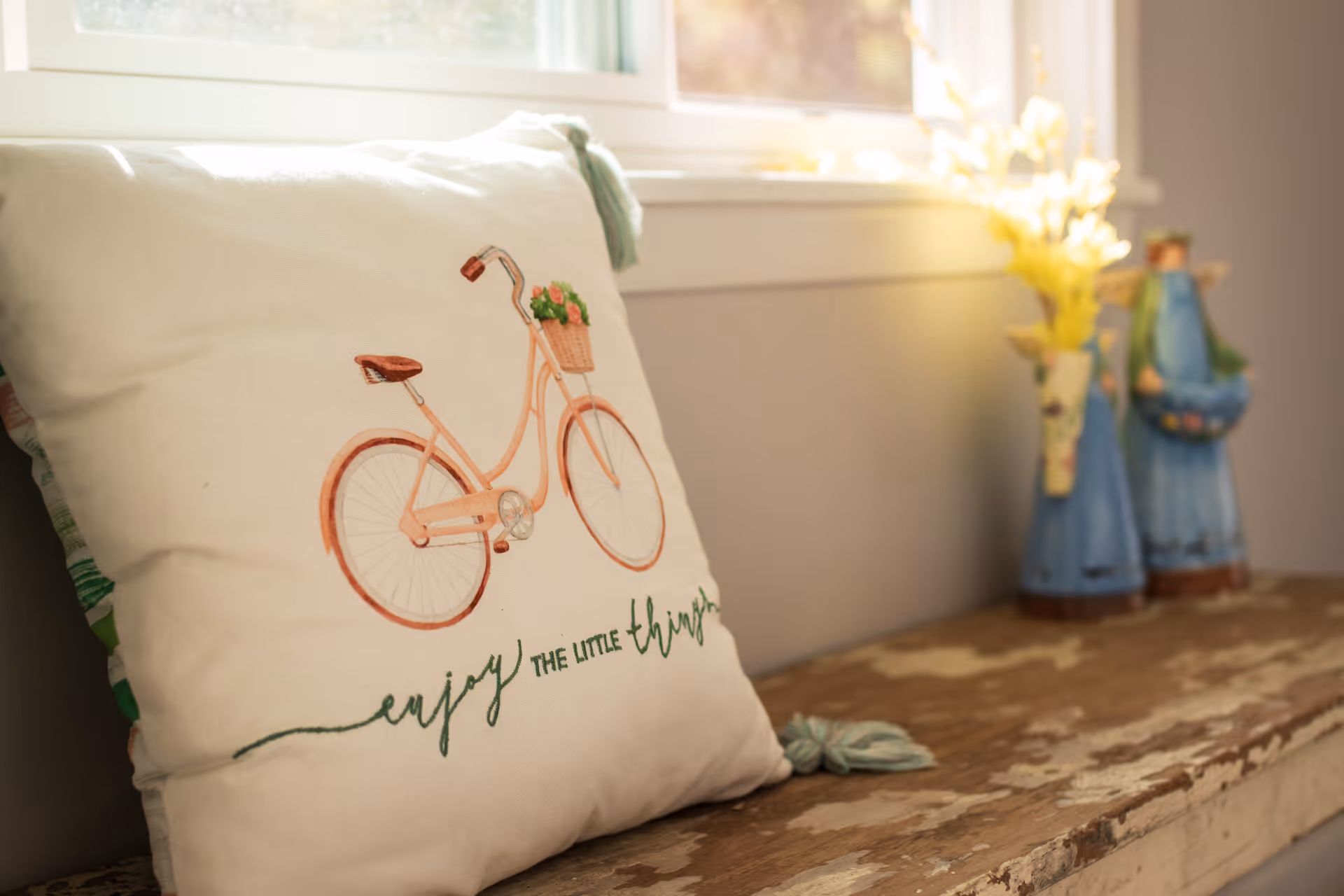 A decorative pillow with a bicycle illustration and the phrase 'enjoy the little things' placed on a rustic wooden bench near a window with sunlight coming through. Two blue vases with yellow flowers are on the bench in the background.