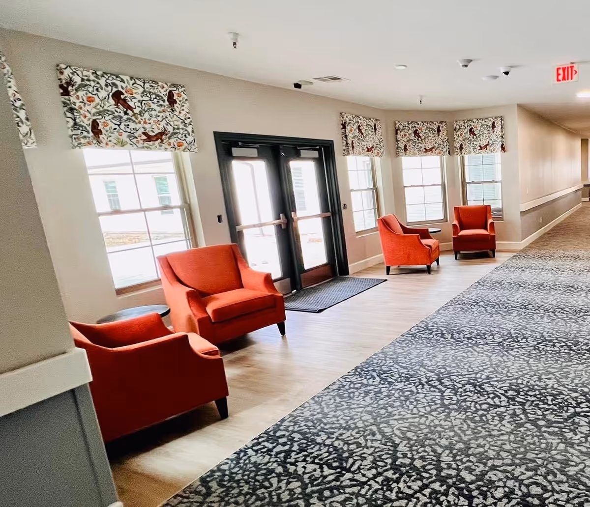 A bright hallway with large windows covered by patterned valances, featuring four orange armchairs arranged along the wall near double glass exit doors. The floor has a combination of light wood and patterned carpet, and there is an exit sign visible on the ceiling.