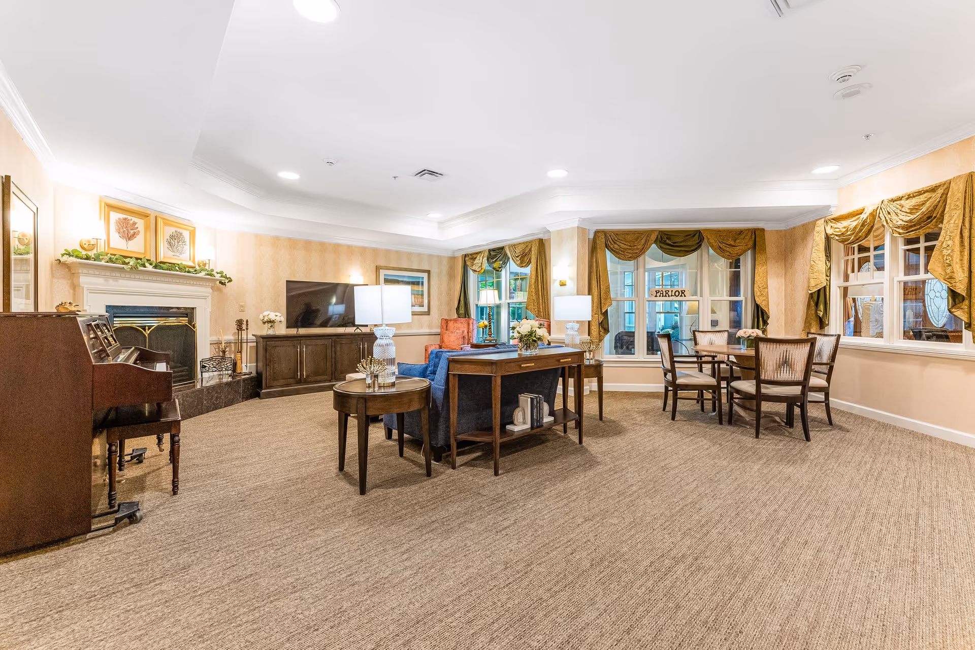A spacious and well-lit living room area in a senior living facility with beige carpet and wallpaper. The room features a fireplace with decorative artwork above it, a piano, a flat-screen TV on a wooden cabinet, a blue sofa with side tables and lamps, and a round table with four chairs near windows adorned with gold curtains. The windows have a sign that reads 'PARLOR'.