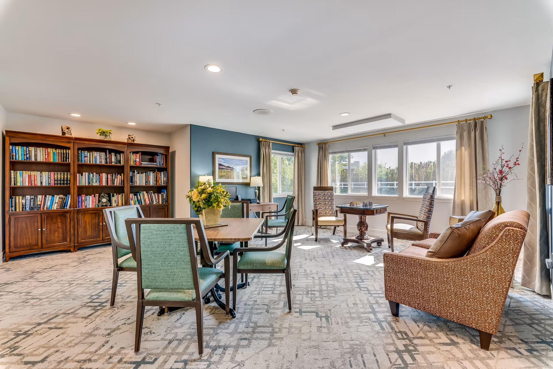 A bright and spacious senior living common area with a large wooden bookshelf filled with books on the left, a table with green cushioned chairs in the center, and a cozy seating area with a patterned sofa and two armchairs around a small table with a chessboard near large windows with beige curtains letting in natural light.