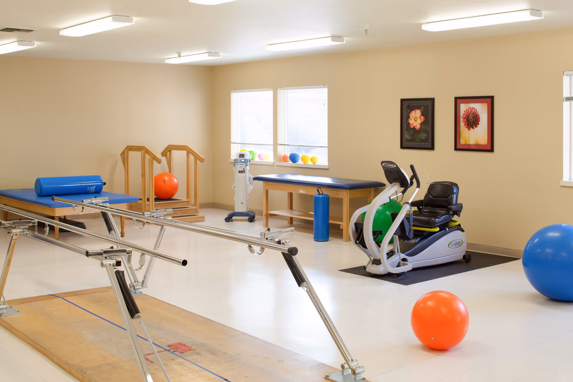 A physical therapy or rehabilitation room with exercise equipment including parallel bars, therapy tables, exercise balls, and a recumbent exercise bike. The room has beige walls, two windows with blinds, and two framed floral pictures on the wall.