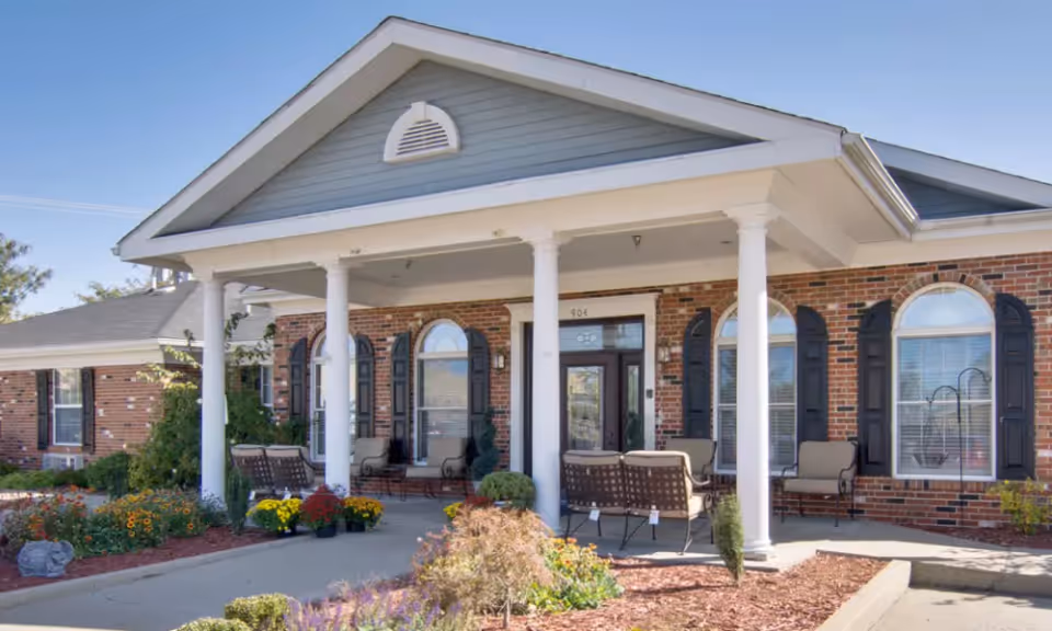 Front exterior view of Westport Estates Senior Living building with a covered porch supported by white columns, outdoor seating, brick walls, arched windows with black shutters, and landscaped garden beds with flowers and shrubs.