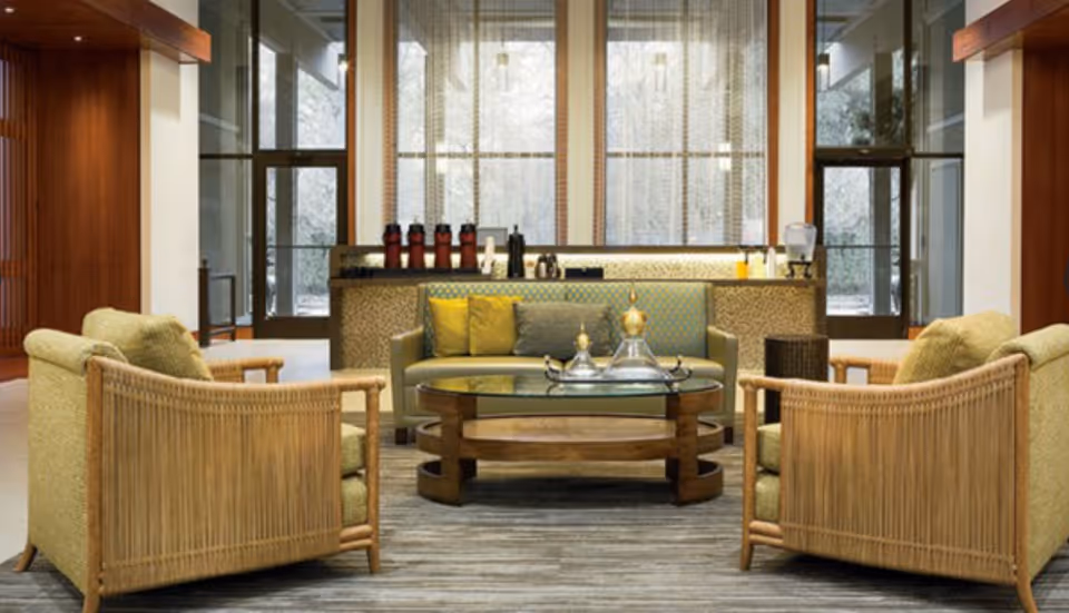 A cozy seating area in a senior living facility with two cushioned armchairs and a cushioned sofa arranged around a wooden coffee table with a glass top. Behind the seating area is a counter with coffee dispensers, cups, and a water dispenser. Large windows with sheer curtains allow natural light to fill the space.