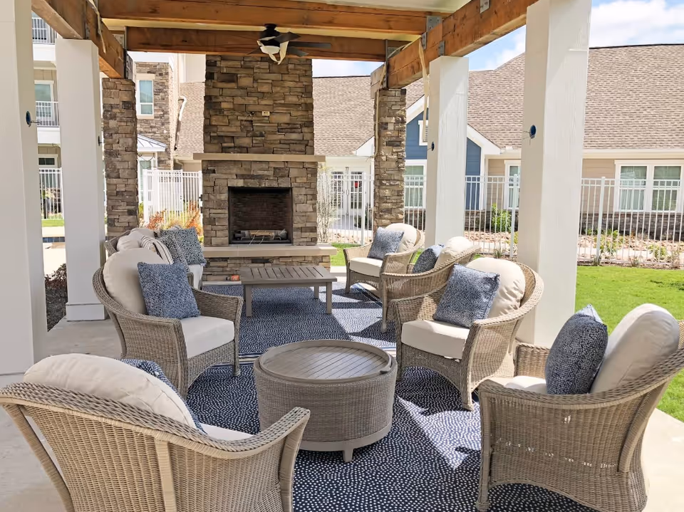 Outdoor covered seating area with wicker chairs and cushions arranged around a coffee table and a stone fireplace, with a ceiling fan overhead and a view of a fenced lawn and residential buildings in the background.