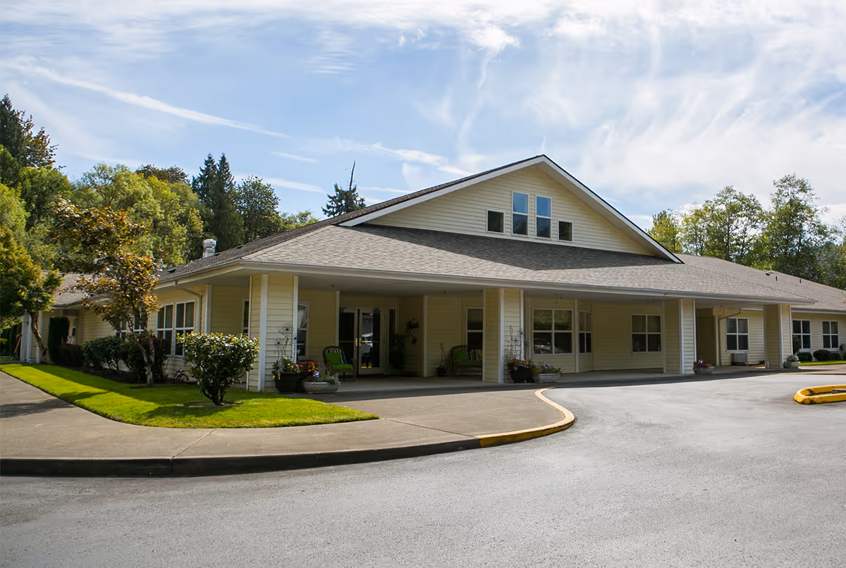 Exterior view of a single-story senior living facility building with a covered entrance, surrounded by green trees and shrubs under a partly cloudy sky.