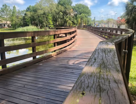 A wooden boardwalk with railings curves gently through a green outdoor area with trees and a pond under a partly cloudy blue sky.