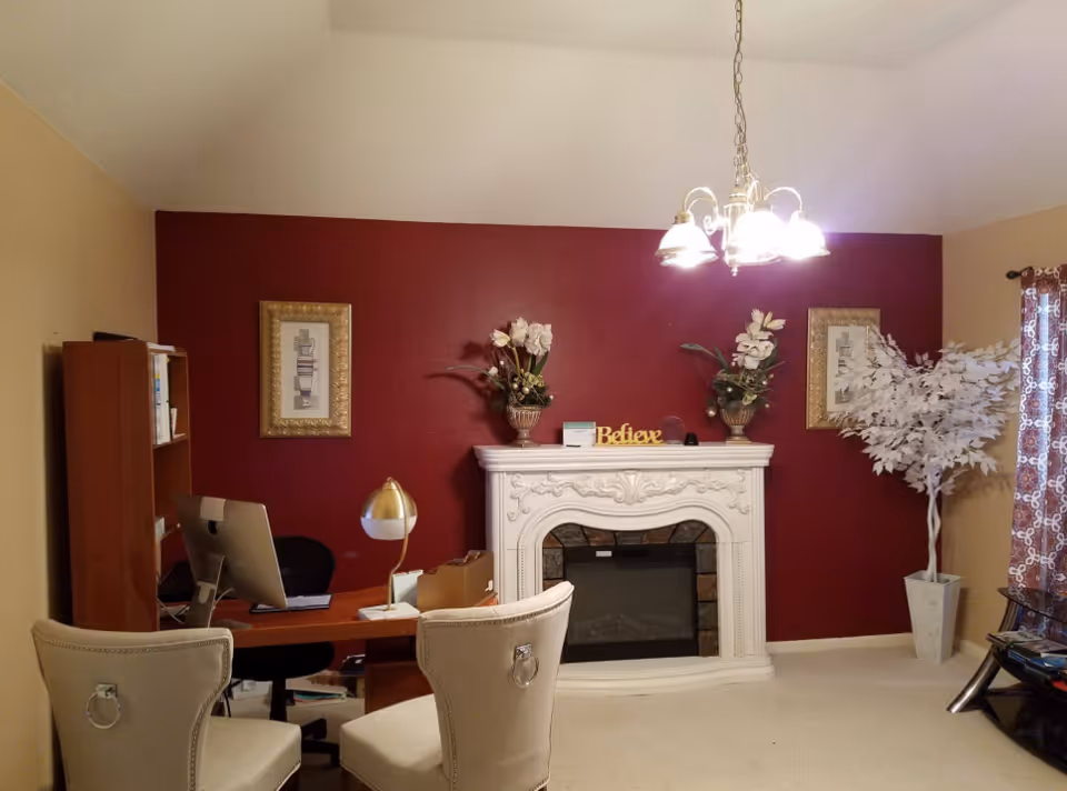 Office-like living room with burgundy accent wall, ornate white fireplace, desk, computer, and chairs.
