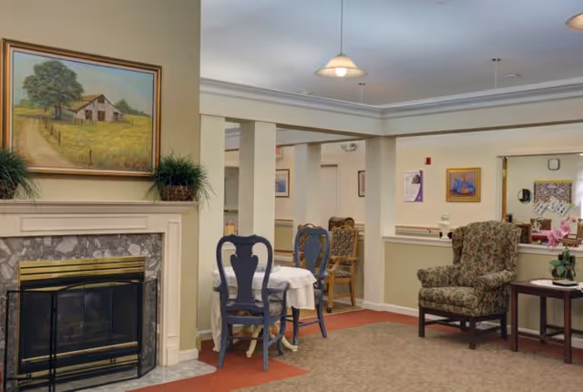 A cozy senior living common area featuring a fireplace with a painting of a barn above it, a small round table with two blue chairs, a patterned armchair, and a side table with a plant. The room has beige walls, carpeted floors, and soft overhead lighting.