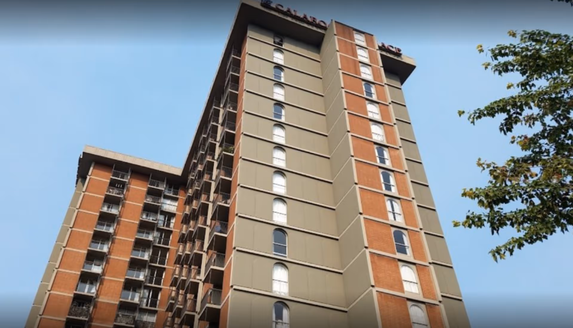 Low-angle view of a tall brick and concrete residential building with balconies and arched windows against a blue sky.