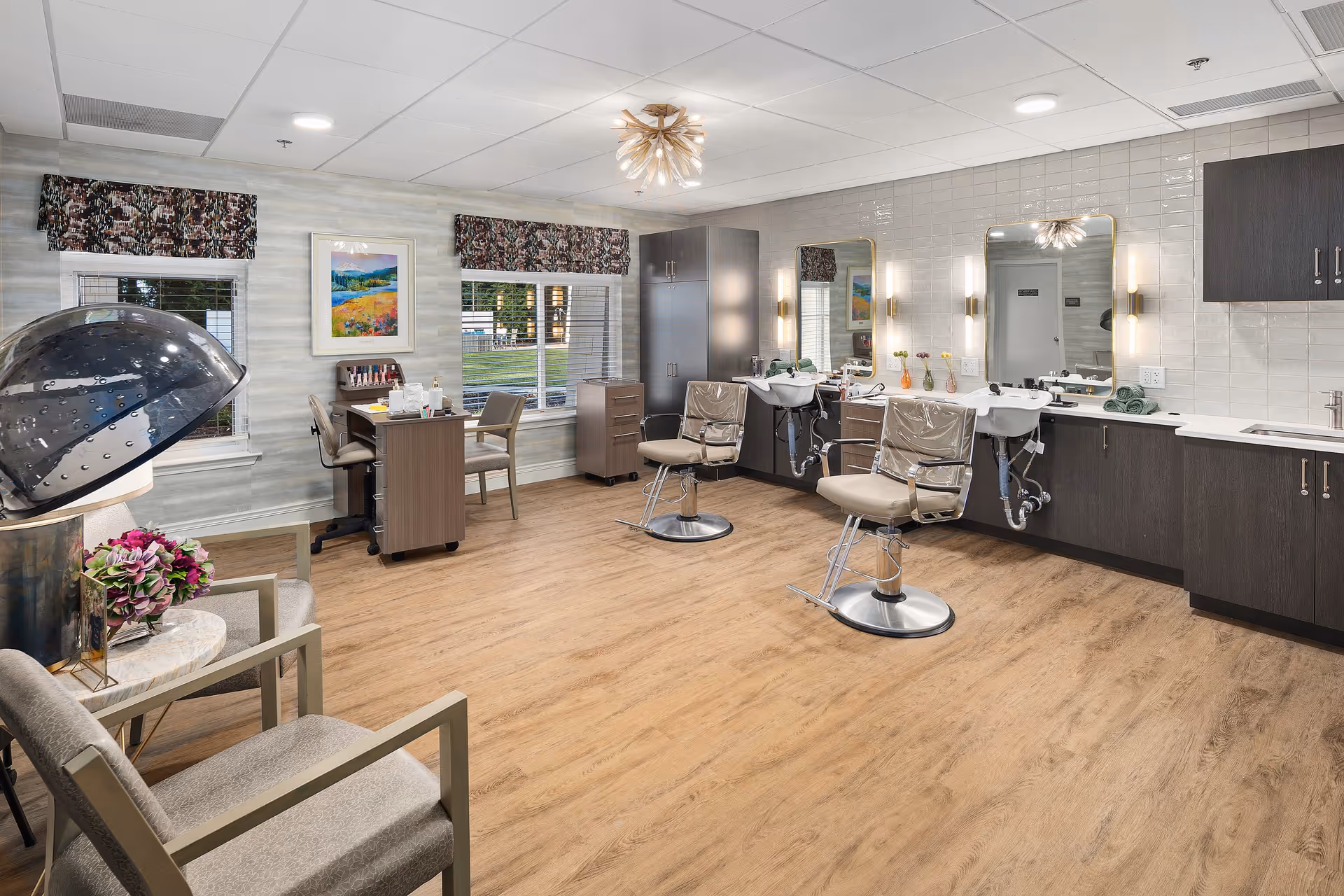 A bright and clean salon area inside a senior living facility with two salon chairs in front of sinks and mirrors, a hair dryer chair, a small desk with chairs near two windows, and modern cabinetry along the wall. The room has wood flooring, floral valances on the windows, and a decorative ceiling light fixture.